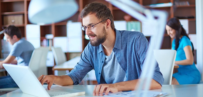 Person working on a laptop with coworkers at desks in the background 